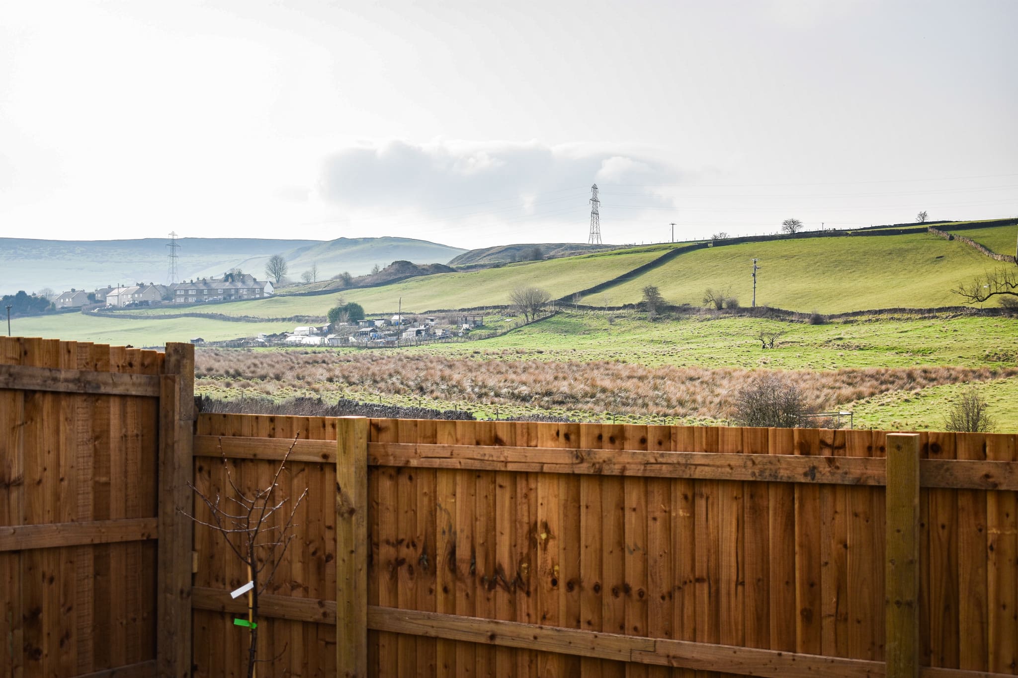 A drystone wall tracing the hillside beyond a Hallstead Meadow garden fence, with the village and a pylon in the distance